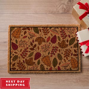 May include: A brown coir doormat with a black border featuring a pattern of colourful autumn leaves and branches.