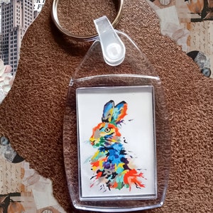 Puede incluir: Llavero acrílico transparente con una pintura abstracta y colorida de un conejo. El conejo presenta tonos vibrantes de azul, naranja, amarillo y verde. El llavero tiene un anillo de metal plateado para llaves.