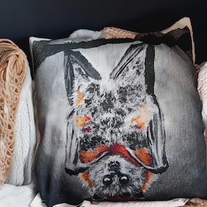 Könnte beinhalten: Quadratisches Zierkissen mit einem stilisierten Fledermaus-Design. Die Fledermaus ist kopfüber dargestellt, mit grau-weißem Körper, schwarzen Flügeln und orangefarbenen und roten Akzenten. Der Hintergrund ist neutral.