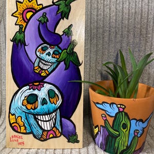 May include: A wooden panel painted with a purple eggplant and a blue skull with yellow flowers. The skull is smiling and has a pink flower on its forehead. The panel is signed "Daniel Luna 2023".
