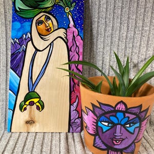 May include: A wooden art piece and a terracotta pot. The art piece features a figure in a white hooded garment against a backdrop of mountains and a starry sky. The pot is decorated with a purple and pink face design, holding a green plant.