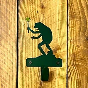 May include: A green metal wall hook featuring a silhouette of a frog holding a wand. The frog is perched on a small platform above the hook. The hook is mounted on a wooden background with vertical wood grain.
