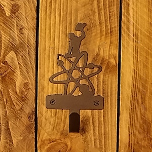May include: A metal wall hook featuring a silhouette of a person atop an atom symbol. The hook is dark brown and mounted on a wooden background with vertical planks. The design is a unique decorative element.