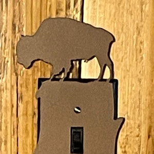 May include: A metal light switch cover with a bison silhouette on top. The cover is brown and has a decorative shape. The switch is in the on position. The background is a wooden wall.