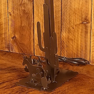 May include: A metal table lamp with a yellow lampshade and a cactus and animal silhouette design. The lamp base features a cactus with a donkey, a bird, and a cow.