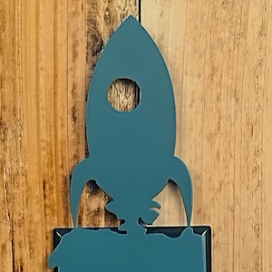 May include: A teal blue light switch cover with a rocket ship design. The switch cover is mounted on a wooden wall.