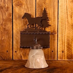 May include: A rustic wall sconce featuring a metal silhouette of horses and trees. The sconce has a rectangular backplate, a decorative metal arm, and a frosted glass bell-shaped shade. The fixture is mounted on a wooden wall.