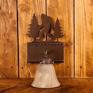 May include: A rustic wall sconce featuring a metal silhouette of Bigfoot walking between two trees. The sconce has a brown finish and a bell-shaped, frosted glass shade. The fixture is mounted on a wooden wall.