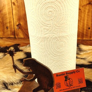May include: A rustic paper towel holder with a silhouette of a frog holding a star wand. The holder is made of brown metal and has a small orange tag that says "Rustic Supply Co". A roll of white paper towels is in the holder.