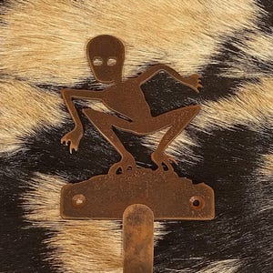 May include: A bronze-colored metal wall hook featuring a stylized figure with a large head and outstretched arms and legs. The hook is mounted on a rectangular plate with two holes for mounting. The background is a pattern of black and tan fur.