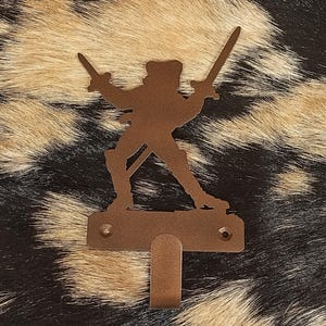 May include: A brown metal wall hook featuring a silhouette of a person holding two swords. The hook has a rectangular base with two holes for mounting and a curved hook below. The background is a mottled pattern of black, brown, and tan.