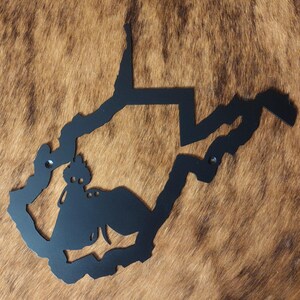 May include: A black metal wall art piece in the shape of the state of West Virginia, mounted on a brown and tan textured background. The artwork features a silhouette of the state's outline with a decorative element.