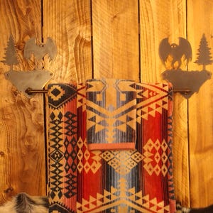May include: A rustic bathroom scene features a patterned towel set in red, black, and tan, hanging on a metal towel rack. The rack is adorned with a bat and tree silhouette design, mounted on a wooden wall.