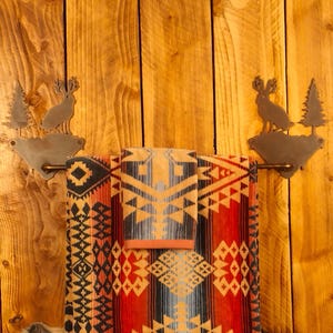 May include: A rustic towel rack with a decorative metal deer and tree design. The rack holds a folded towel set with a geometric pattern in red, black, blue, and tan. The background is a wooden wall.