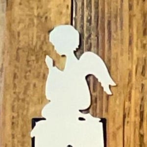 May include: A white angel-shaped light switch cover is mounted on a wooden surface. The cover has a cutout for the switch and is adorned with an angel figure at the top. The wooden surface has a natural wood grain.