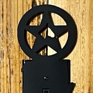 May include: Black metal light switch cover with a pentagram design. The switch cover is rectangular with a decorative border and a toggle switch. The pentagram is within a circular frame above the switch.