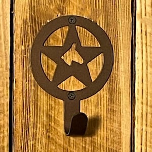 May include: A rustic, brown metal wall hook featuring a star within a circle design. The hook is mounted on a wooden surface with visible grain and knots. The star has a cutout of a state shape.