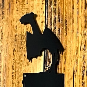 May include: Black metal light switch cover with a dragon silhouette design. The dragon is perched above the switch plate. The background is a wooden surface with vertical grain.