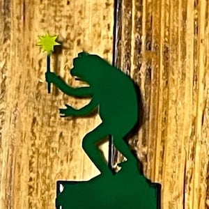 May include: A green metal light switch cover featuring a frog holding a wand with a star on top. The frog is standing on top of the switch plate. The switch plate is rectangular with a black border.