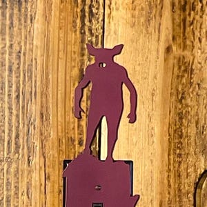 May include: A burgundy metal light switch cover featuring a silhouette of a creature with horns, standing on top of the switch plate. The switch plate is black with a toggle switch and screw details. The background is a wooden wall.