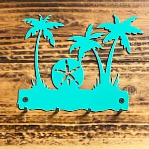 May include: Teal metal key holder shaped like a beach scene with palm trees and a sand dollar. The key holder has four hooks for hanging keys.