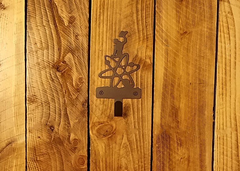 May include: A metal wall hook featuring a silhouette of a person atop an atom symbol. The hook is dark brown and mounted on a wooden background with vertical planks. The design is a unique decorative element.