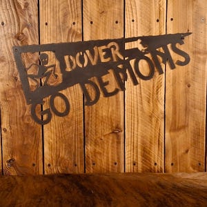 May include: A dark metal sign with the words "DOVER DEMONS" cut out. The sign also features a cutout of a small alien figure. The sign is mounted on a wooden background.