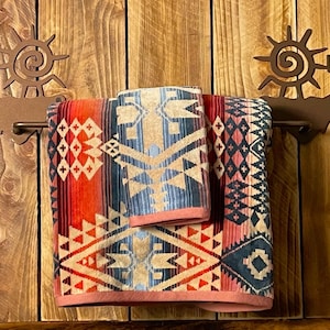 May include: A rustic towel rack with a southwestern design. The rack features two metal sun and cactus figures with a towel bar in the middle. Two towels with a geometric pattern are hanging on the rack.