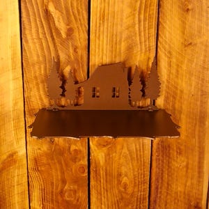 May include: A brown metal shelf with a house and tree silhouette design. The shelf is mounted on a wooden wall with vertical planks. The house has two windows and is flanked by stylized trees.