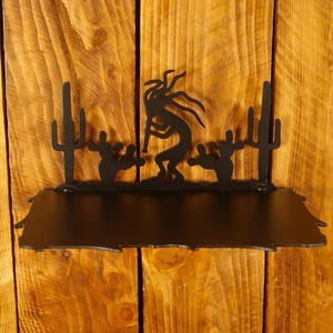 May include: Black metal shelf featuring a Kokopelli figure playing a flute, flanked by two cacti. The shelf is mounted on a wooden plank wall, showcasing a Southwestern theme.