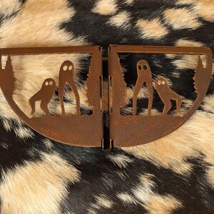 May include: A rusty metal art piece featuring two halves of a semi-circular design. Each half depicts a stylized forest scene with two ghost-like figures. The artwork is set against a backdrop of a black, brown, and tan animal hide.