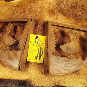 May include: Two brown metal shelf brackets featuring a cutout silhouette of a duck wearing a top hat. A yellow rectangular card is placed between the brackets with the text The Rustic Supply Co, With Quackiness we will prevail, and a cactus.