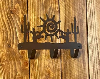 May include: A metal wall hook with a southwestern design featuring a sun, cacti, and two figures. The hook has three hooks for hanging items.