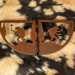 May include: Two rusty metal bookends, each featuring a silhouette of a creature holding a star-shaped object. The bookends have a curved design with a forest scene. The background is a cowhide pattern.