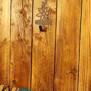 May include: A metal wall hook with an atom design, mounted on a wooden plank background. The hook is dark-colored and has a single hook for hanging items. The wood has a warm, natural tone with visible grain.