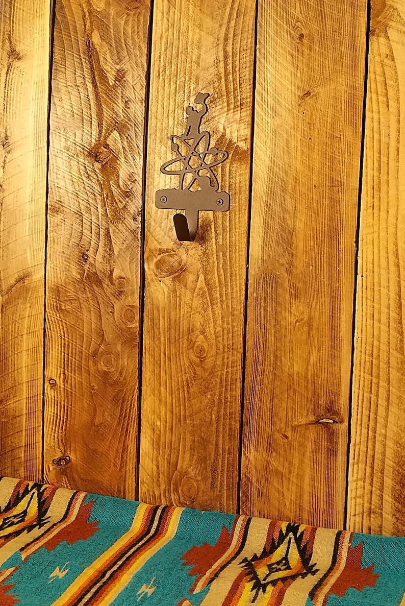 May include: A metal wall hook with an atom design, mounted on a wooden plank background. The hook is dark-colored and has a single hook for hanging items. The wood has a warm, natural tone with visible grain.