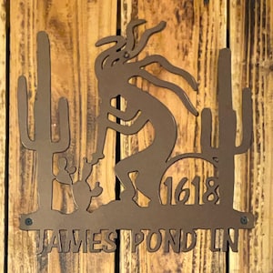 May include: A brown metal sign featuring a Kokopelli figure playing a flute, with two cacti on either side. The sign includes the number 1618 and the text "JAMES POND LN".