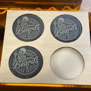 Puede incluir: Un juego de cuatro posavasos de pizarra negra con un diseño blanco. Los posavasos presentan un esqueleto con un signo de paz y el texto "Dads with Beards are Better".