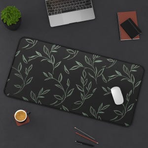 May include: A black desk mat with a repeating pattern of light green leaves and vines. A white computer mouse sits on the mat, along with a laptop, a small potted plant, a cup of coffee, and stationery.