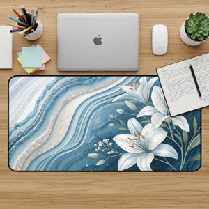 May include: A rectangular desk mat featuring a watercolor design with blue, white, and beige abstract patterns. White lilies with green leaves are on the right side of the mat. The mat is on a wooden desk with a laptop, mouse, and other office supplies.
