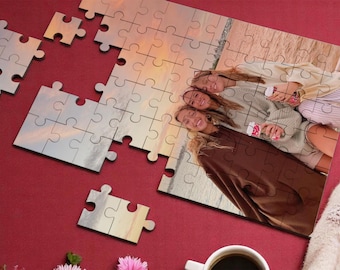 May include: A jigsaw puzzle with a photo of three friends smiling at the camera. The puzzle is partially completed, with some pieces missing. The background of the photo is a beach with a sunset.