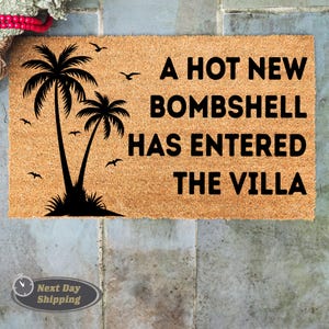May include: A rectangular tan coir doormat featuring a black graphic of two palm trees and several birds in flight. The text A HOT NEW BOMBSHELL HAS ENTERED THE VILLA is printed in bold black capital letters on the right side of the mat surface.