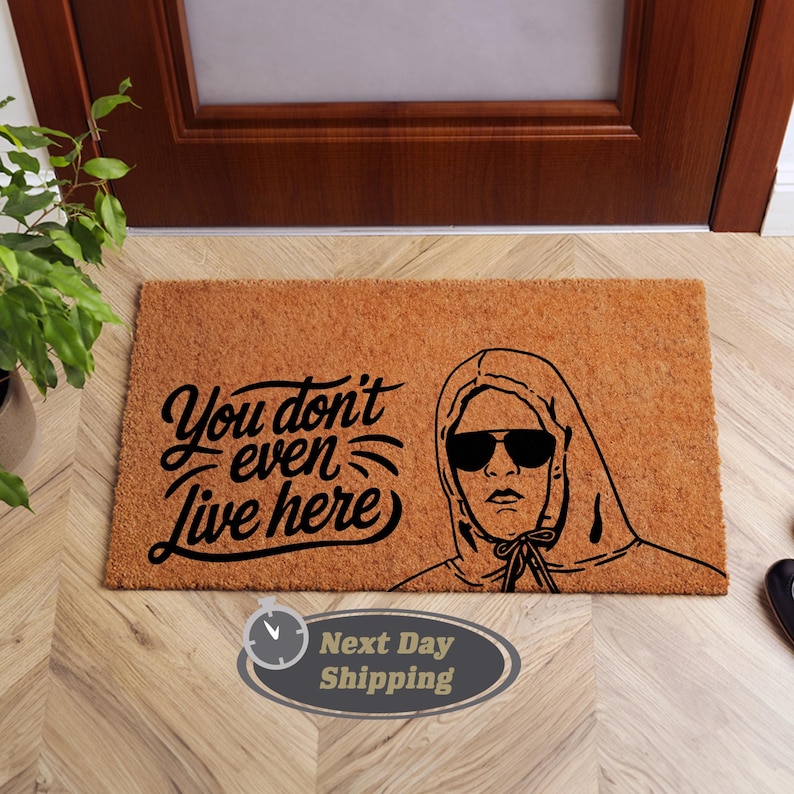 Pode incluir: Um tapete de coco castanho com o texto "You don't even live here" em fonte cursiva preta. Uma ilustra&ccedil;&atilde;o em preto e branco de uma pessoa usando um moletom com capuz e &oacute;culos de sol est&aacute; no lado direito do tapete.