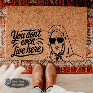 Pode incluir: Um tapete de coco castanho com o texto "You don't even live here" em fonte cursiva preta. Uma ilustra&ccedil;&atilde;o em preto e branco de uma pessoa usando um moletom com capuz e &oacute;culos de sol est&aacute; no lado direito do tapete.