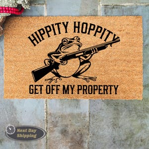 May include: A rectangular doormat made of natural coir fiber. The mat features a black illustration of a frog holding a shotgun, with the text "HIPPITY HOPPITY GET OFF MY PROPERTY" in black, arched above and below the frog.