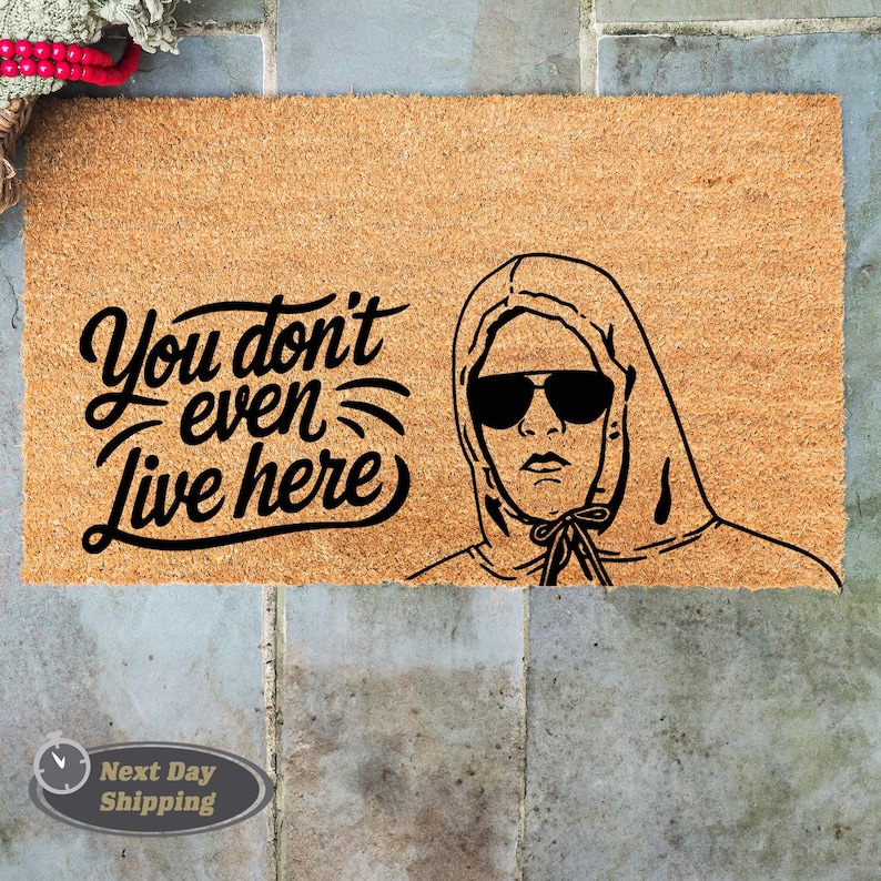 Pode incluir: Um tapete de coco castanho com o texto "You don't even live here" em fonte cursiva preta. Uma ilustra&ccedil;&atilde;o em preto e branco de uma pessoa usando um moletom com capuz e &oacute;culos de sol est&aacute; no lado direito do tapete.