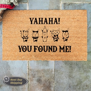 May include: A brown coir doormat with the text "YAHAHA! YOU FOUND ME!" in black. The text is surrounded by five cartoon characters, four of which are wearing striped shirts and hats, and one is a long, thin, green character.