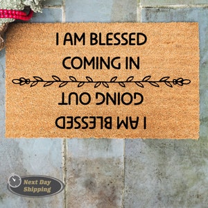 May include: A brown coir doormat with the text "I am blessed coming in I am blessed going out" in black lettering. A decorative vine design separates the two lines of text.