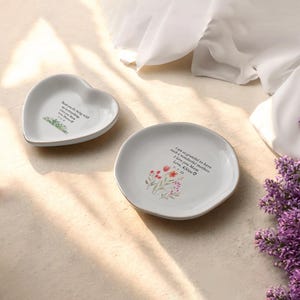 Puede incluir: Dos platos de cer&aacute;mica blanca para joyas con ribete dorado. Uno de los platos tiene forma de coraz&oacute;n y el otro es redondo. Ambos platos presentan dise&ntilde;os florales y mensajes escritos a mano. El plato en forma de coraz&oacute;n dice "Gracias por estar conmigo en todo. Te quiero mam&aacute;. Con amor, [Nombre] 8.18.23". El plato redondo dice "Estoy tan agradecida de tener una madre tan maravillosa. Te quiero mam&aacute;. Con amor, Khloe 7.7.23".