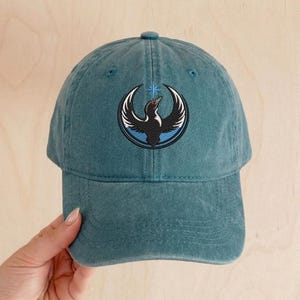 May include: Teal baseball cap with a curved brim. The cap features an embroidered emblem of a black bird with white wings, a red eye, and a blue and white star. The cap is made of a soft, textured fabric.